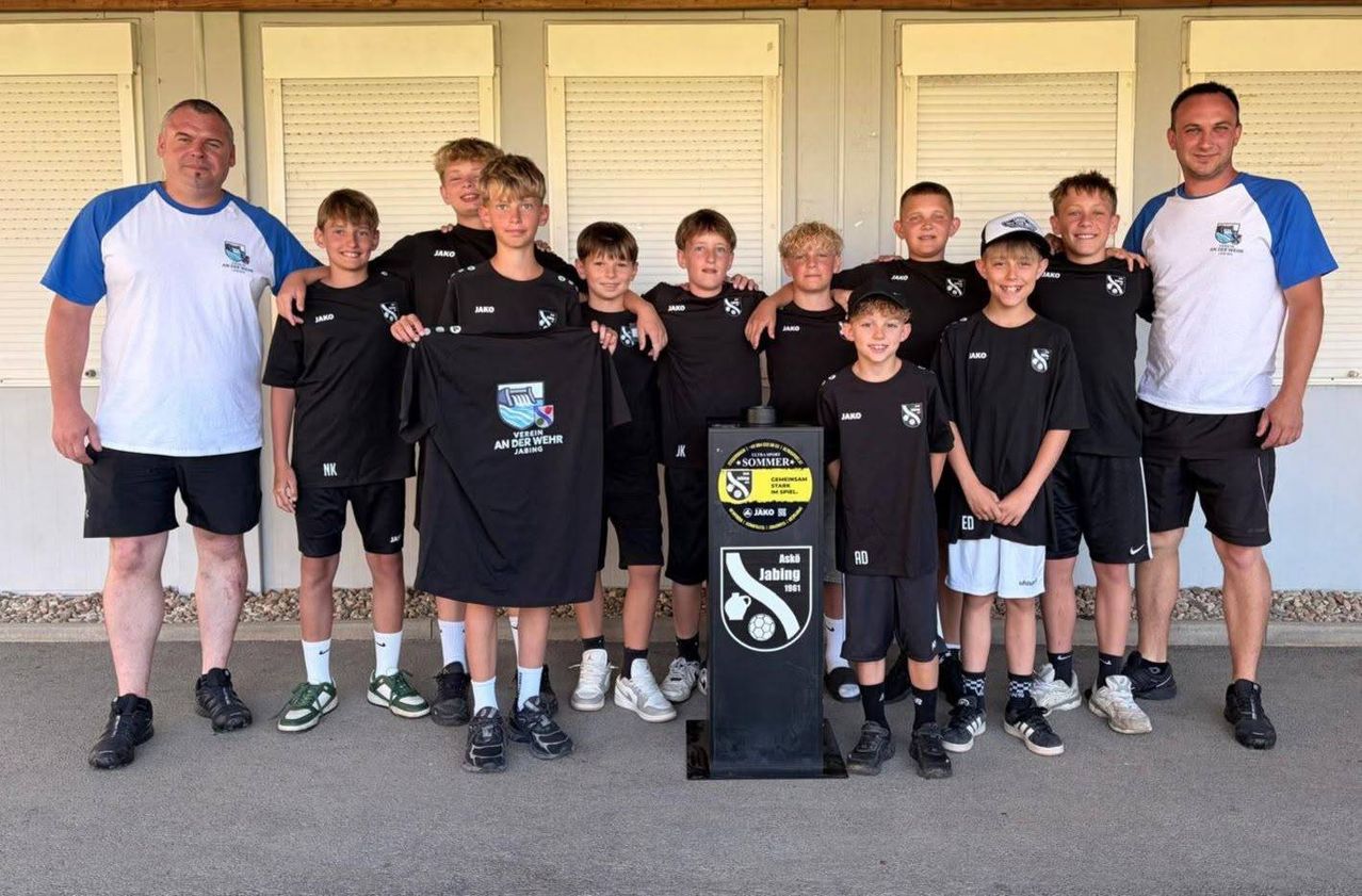 Eine Gruppe junger Jungen in schwarzer und weißer Sportbekleidung posiert mit einem Pokal für ein Foto. Sie stehen vor einer weißen Wand mit Jalousien.