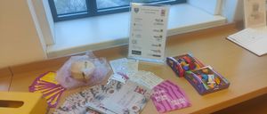 Ein Tisch mit verschiedenen Broschüren, einer herzförmigen Box und zwei Schachteln mit Snacks vor einem Fenster. Das Fenster bietet einen Blick auf geparkte Autos und Gebäude.