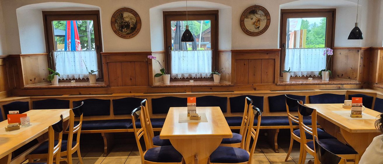 Ein Innenrestaurant mit einem Holztisch und Stühlen. Der Tisch hat eine Speisekarte darauf. Der Raum hat ein Fenster mit einem weißen Vorhang und Topfpflanzen auf der Fensterbank. Der Raum hat Holzbänke und Wände.