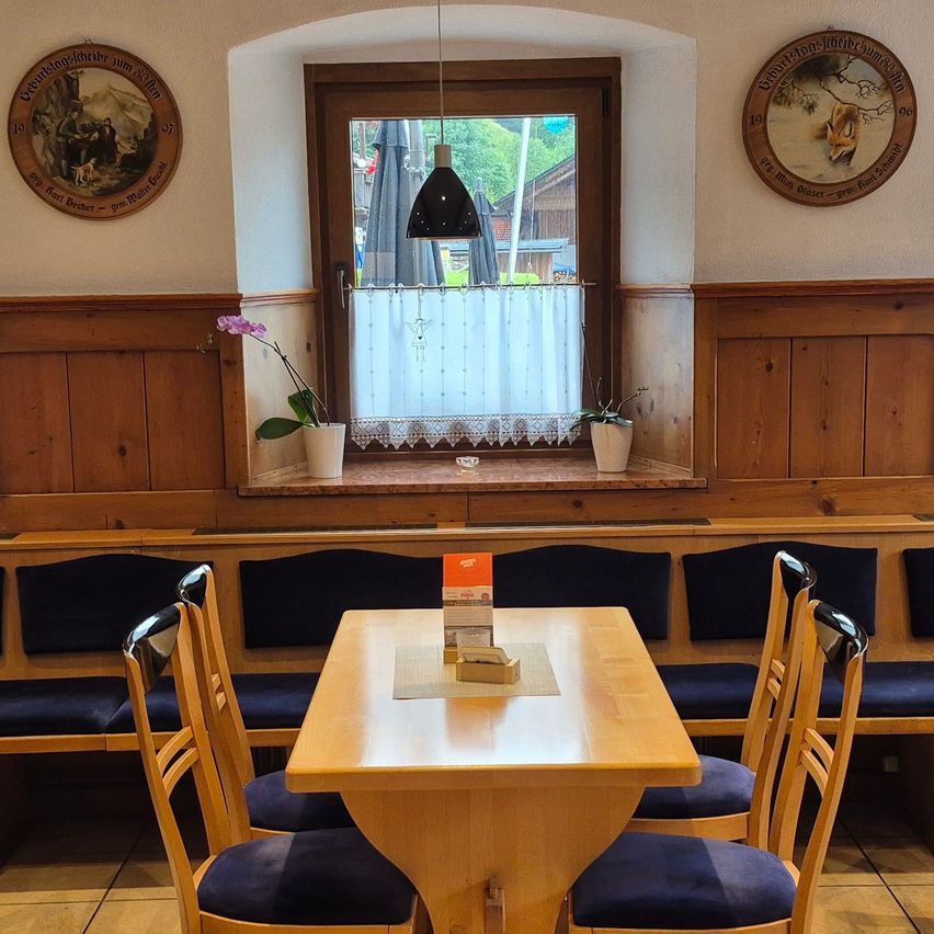 Ein Innenrestaurant mit einem Holztisch und Stühlen. Der Tisch hat eine Speisekarte darauf. Der Raum hat ein Fenster mit einem weißen Vorhang und Topfpflanzen auf der Fensterbank. Der Raum hat Holzbänke und Wände.