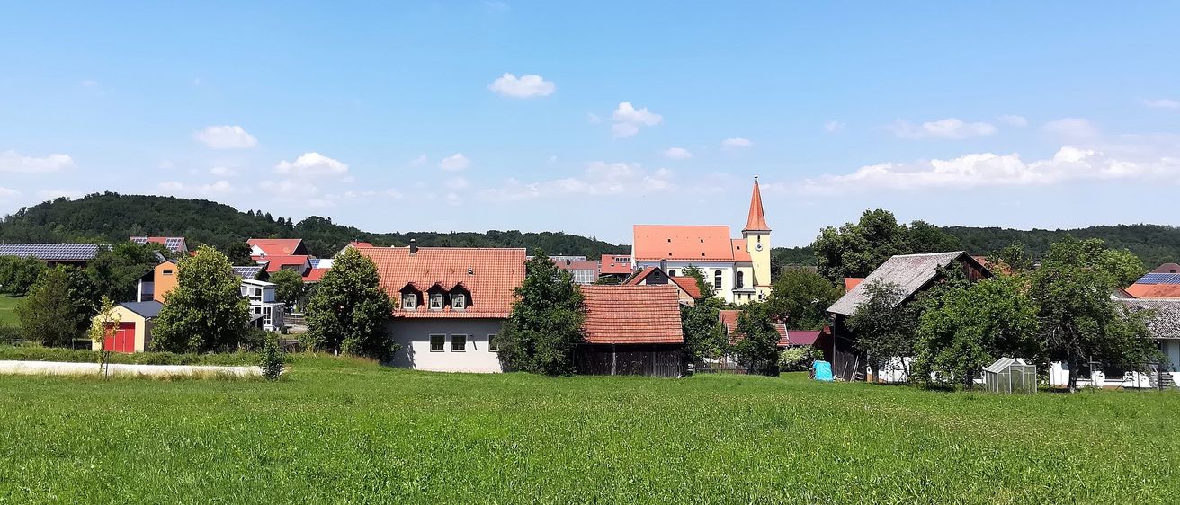 Ein Dorf mit rot gedeckten Häusern und einer Kirche unter blauem Himmel. Das Dorf ist von üppigen grünen Feldern und Bäumen umgeben.