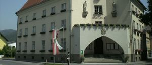 Das Gebäude ist ein Rathaus mit einer Uhr, Statuen und einer rot-weißen Flagge. Der Eingang hat Stufen und einen Balkon mit Blumen.