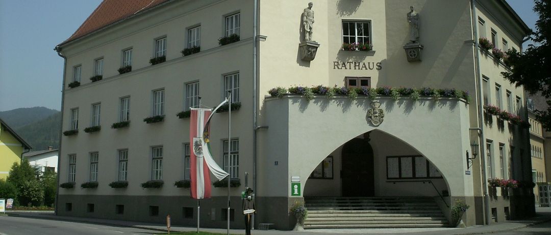 Das Gebäude ist ein Rathaus mit einer Uhr, Statuen und einer rot-weißen Flagge. Der Eingang hat Stufen und einen Balkon mit Blumen.
