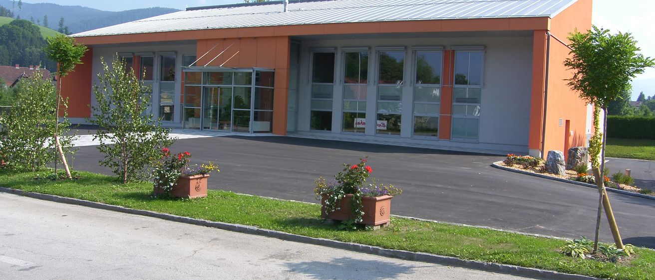 Ein modernes Bürogebäude mit Glasfenstern und orangefarbenen Akzenten, flankiert von zwei Blumenbeeten an einem sonnigen Tag.