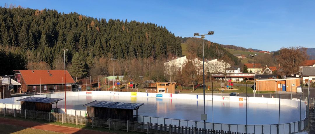 Eine Eislaufbahn im Freien ist von einem weißen Zaun umgeben. Zwei kleine Holzhütten sind auf der Bahn. Straßenlaternen stehen auf beiden Seiten. In der Ferne ein Berg und Häuser.