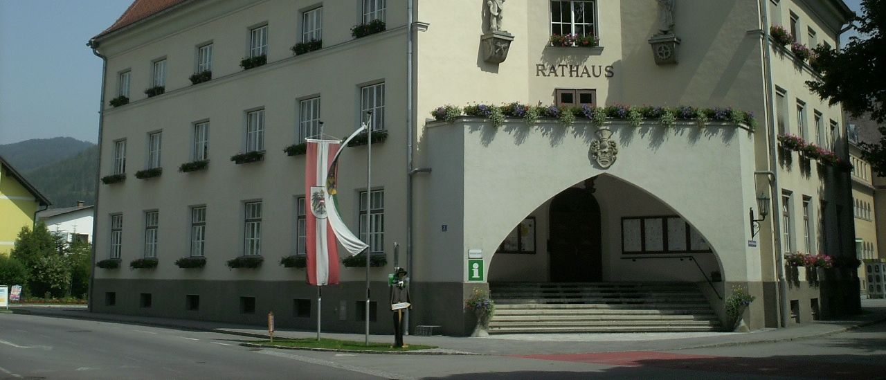 Ein großes weißes Gebäude mit einem braunen Dach und einem Uhrturm, mit Statuen und einem Schild, das Rathaus steht. Es hat viele Fenster, eine Flagge und Pflanzen.