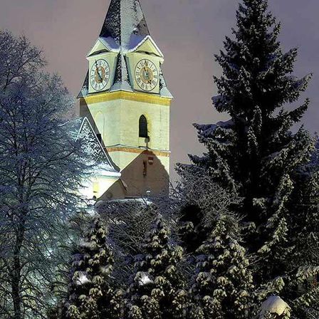 Ein Kirchturm mit zwei Uhren, umgeben von schneebedeckten Bäumen unter einem blauen Nachthimmel.