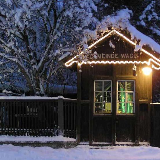 Ein kleines Holzhaus ist nachts von Lichtern beleuchtet und mit Schnee bedeckt. Es trägt ein Schild mit der Aufschrift 'Meine Wache'. Das Haus ist eingezäunt und mit Schnee bedeckt.