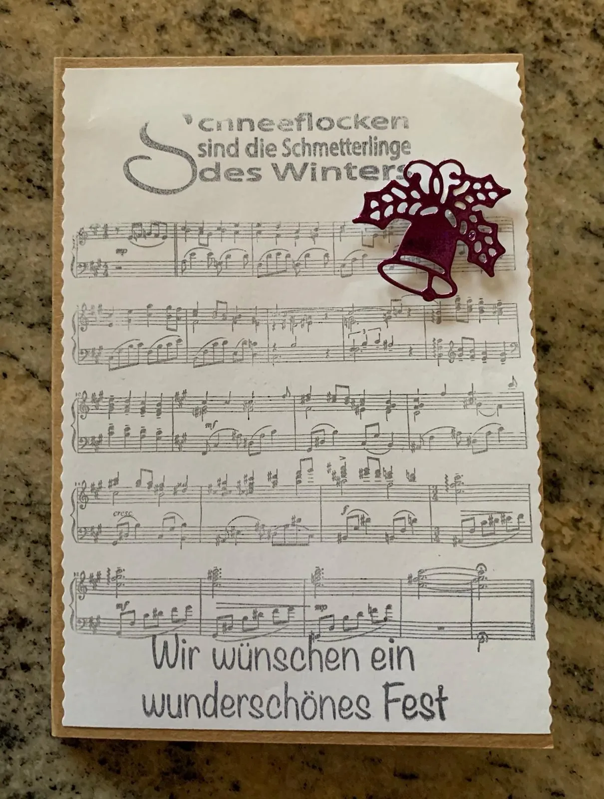 Ein weißes Papier mit Noten und einem deutschen Zitat oben, begleitet von einer dekorativen roten Glocke, liegt auf einer Marmoroberfläche.
