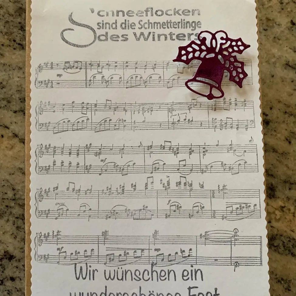 Ein weißes Papier mit Noten und einem deutschen Zitat oben, begleitet von einer dekorativen roten Glocke, liegt auf einer Marmoroberfläche.