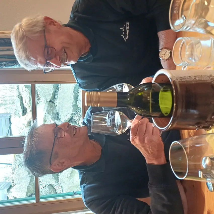 Zwei Männer trinken Wein aus einer Flasche, die in einem Eimer steht. Einer schenkt ein, während der andere trinkt. Sie tragen schwarze Hemden mit Logos und Brillen. Gläser stehen auf dem Tisch, und ein Fenster zeigt eine Felswand.