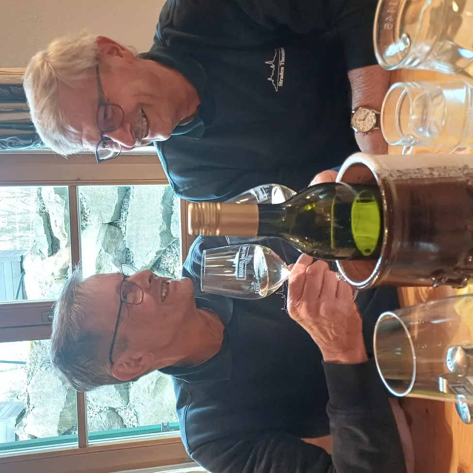 Zwei Männer trinken Wein aus einer Flasche, die in einem Eimer steht. Einer schenkt ein, während der andere trinkt. Sie tragen schwarze Hemden mit Logos und Brillen. Gläser stehen auf dem Tisch, und ein Fenster zeigt eine Felswand.