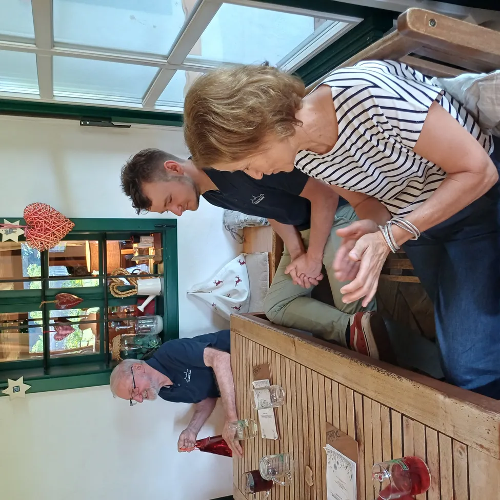 Drei Menschen sind in einem Raum. Eine Frau steht auf einem Holzregal, während ein Mann eine Flasche hält und ein anderer Mann hockt. Der Raum hat ein Holzregal, ein Fenster mit grünem Rahmen und eine Glastür mit einer Reflexion des Raums.