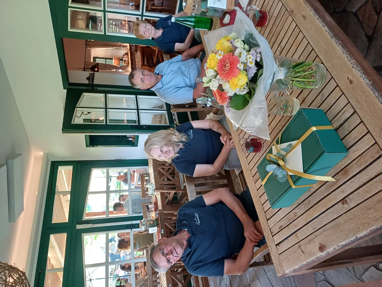 Eine Gruppe von Menschen sitzt an einem Tisch mit einem Blumenstrauß und einem Geschenkkarton. Sie sind in einem Restaurant mit Glasfenstern und Holzstühlen. Der Raum hat eine warme und gemütliche Atmosphäre.