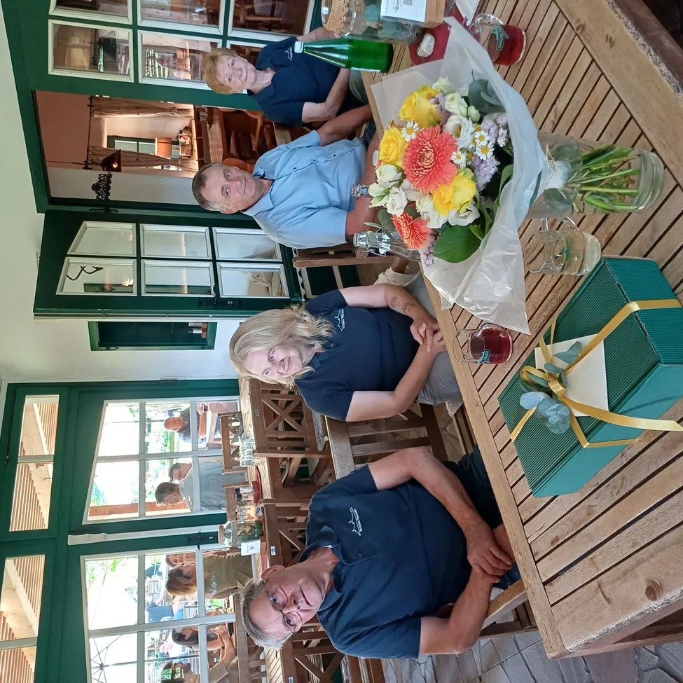 Eine Gruppe von Menschen sitzt an einem Tisch mit einem Blumenstrauß und einem Geschenkkarton. Sie sind in einem Restaurant mit Glasfenstern und Holzstühlen. Der Raum hat eine warme und gemütliche Atmosphäre.