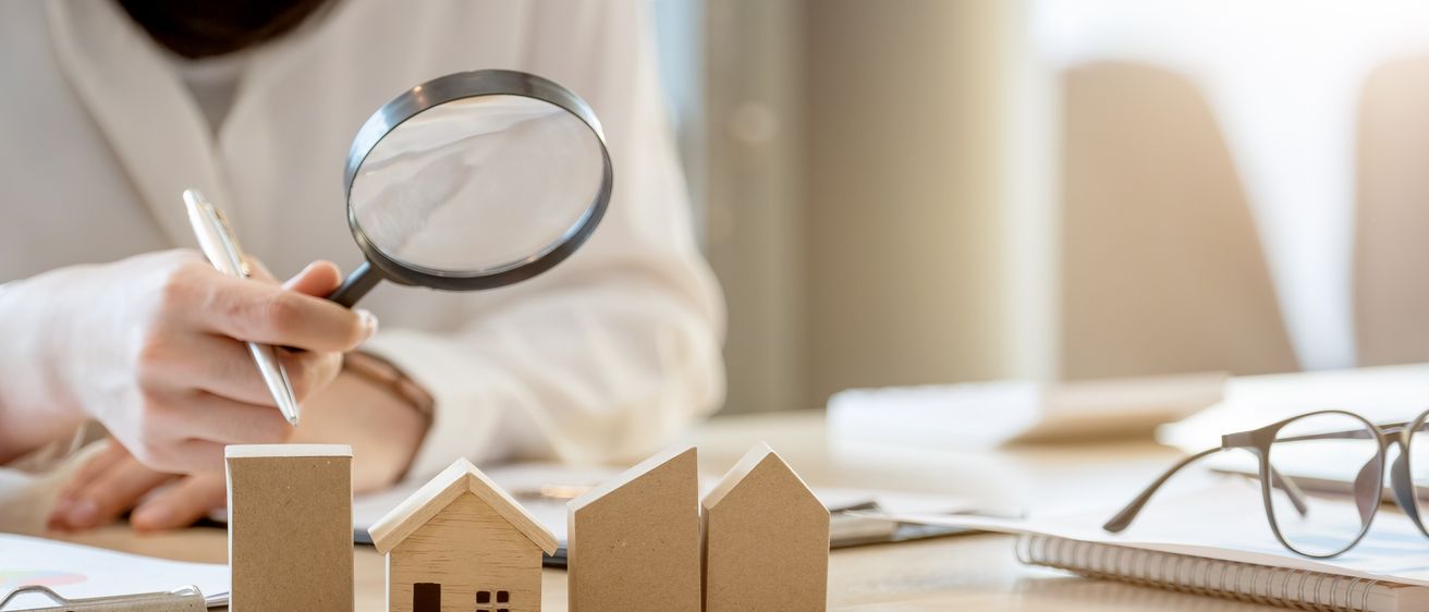 Eine Person untersucht ein Miniaturhausmodell mit einer Lupe, während ein weiteres Hausmodell und Papiere auf dem Tisch liegen.