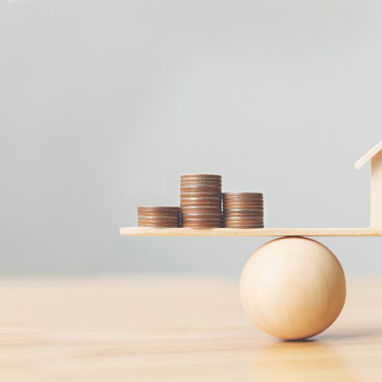 Ein hölzerne Haus balanciert auf einem Wippspiel mit Münzen, die am gegenüberliegenden Ende gestapelt sind. Die Münzen sind in zwei Stapeln angeordnet, einer höher als der andere.