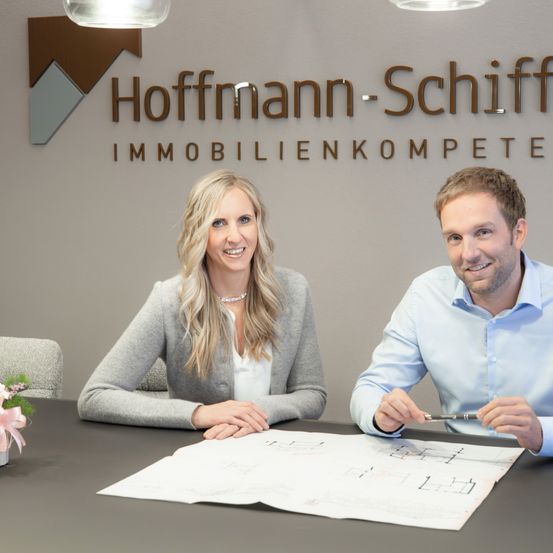 Eine Frau und ein Mann sitzen lächelnd an einem Schreibtisch mit einer Bauzeichnung davor. Hinter ihnen zeigt eine Wand das Hoffmann-Schiff-Logo und den Text Immobilienkompetent.