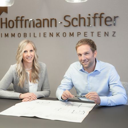 Eine Frau und ein Mann sitzen lächelnd an einem Tisch. Hinter ihnen befindet sich eine Wand mit dem Hoffmann-Schiff-Logo. Die Frau trägt eine graue Strickjacke, der Mann ein blaues Hemd. Sie betrachten Baupläne auf dem Tisch.