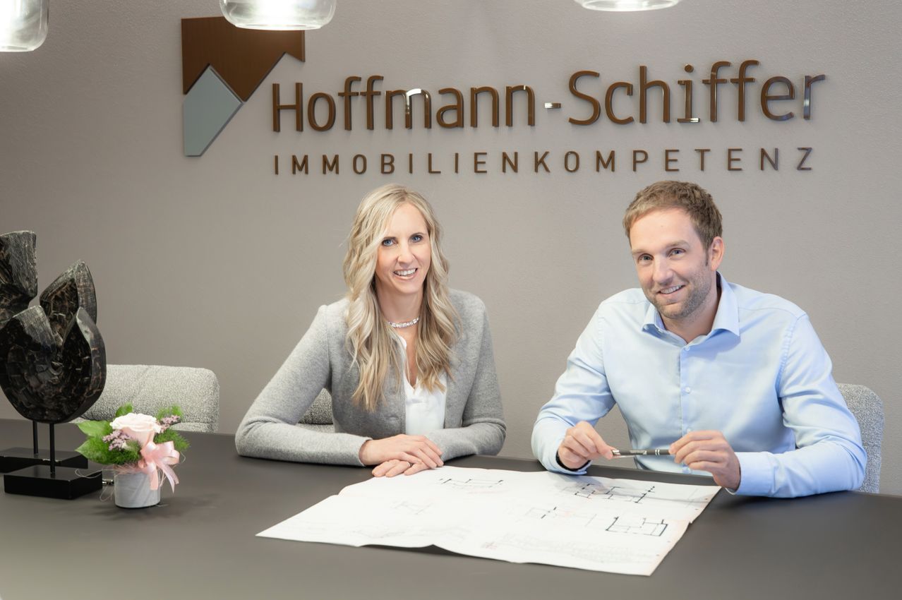 Zwei Fachleute, eine Frau und ein Mann, sitzen lächelnd an einem Tisch. Hinter ihnen befindet sich eine Wand mit dem Hoffmann-Schiff-Logo und Text. Papiere und ein Stift liegen auf dem Tisch.