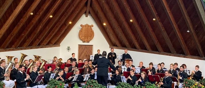 Ein Orchester spielt in einem dreieckigen Veranstaltungsort mit Holzdächern. Zuschauer sitzen vor der Bühne auf dem Rasen. Die Bühne hat viele Pflanzen und Blumen.