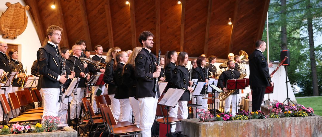 Ein Orchester in Uniform steht mit Musikinstrumenten auf einer Bühne unter einem Holzdach. Sie scheinen sich auf eine Aufführung vorzubereiten.