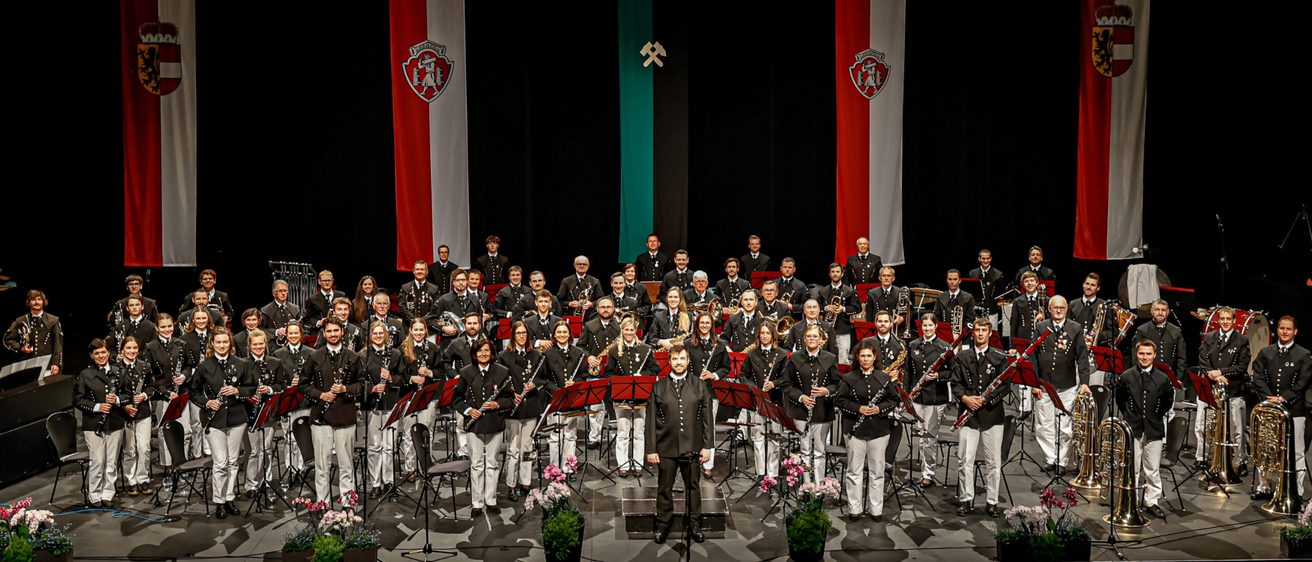 Ein großes Orchester ist auf der Bühne, mit einer Vielzahl von Instrumenten und Uniformen. Dahinter befinden sich drei Banner, von denen eines eine rot-weiße Flagge hat.