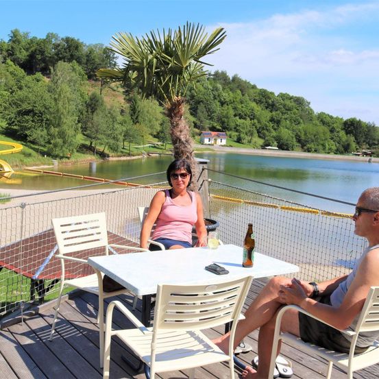 Ein Mann und eine Frau sitzen an einem Tisch auf einer Terrasse mit Blick auf einen See. Dahinter steht eine große Palme, ein Zaun und eine gelbe Rutsche. Der See ist von Bäumen umgeben.