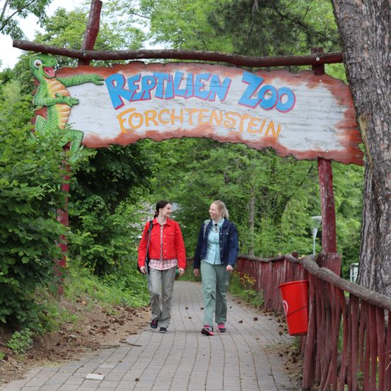 Zwei Frauen gehen an einem Schild für den Reptilienzoo Forchtenstein vorbei, unter dem ein roter Eimer hängt.