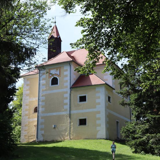 Ein gelbes Gebäude mit einem roten Dach und einem Turm steht auf einem Hügel, umgeben von Bäumen. Eine Person geht auf dem Gras vor dem Gebäude.