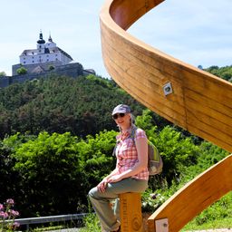 Eine Frau mit Rucksack sitzt auf einer Holzskulptur mit einer Burg auf einem Hügel im Hintergrund.