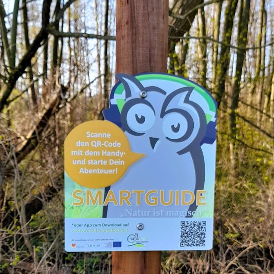 Ein Schild auf einem Holzpfosten sagt 'Scanne den QR-Code mit dem Handy und starte Dein Abenteuer!' Es zeigt eine animierte Eule, den Text 'SMARTGUIDE' und einen QR-Code zum Herunterladen einer App von www.rosalia-kogelberg.at.