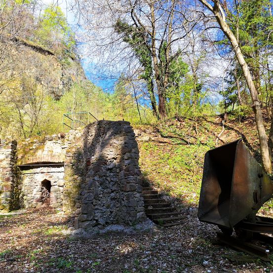 Ein altes Steingebäude mit Moos und einem kleinen Bogeneingang, umgeben von Bäumen und Stufen. Ein rostiger alter Bergbaukübel liegt in der Nähe.