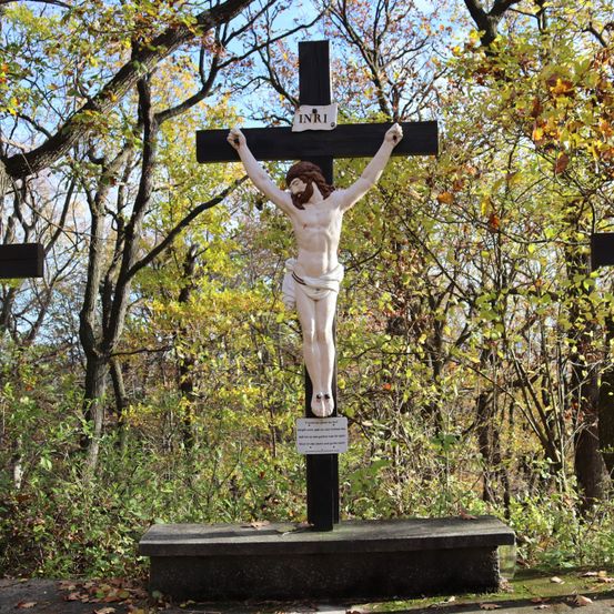 Ein Kruzifix steht prominent in einem Wald, umgeben von Bäumen mit gelben Blättern. Der Kreuz trägt die Inschrift INRI über der Figur Christi.