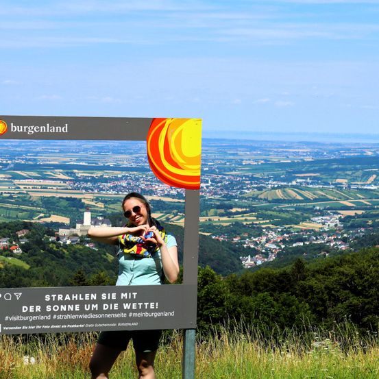 Eine Frau posiert vor einem Schild mit einer malerischen Aussicht auf Burgenland. Das Schild sagt Strahlen Sie mit der Sonne um die Wette! Der Hintergrund zeigt eine Landschaft mit sanften Hügeln, Städten und einer Burg.