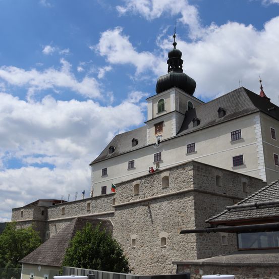 Eine Burg mit einem hohen Turm und einer Kuppel unter einem blauen Himmel mit Wolken. Mehrere Flaggen hängen an der Burgmauer.