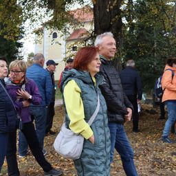 Eine Gruppe von Menschen versammelt sich im Herbst draußen, lächelt und unterhält sich. Eine Frau in einem gelben Pullover steht mit einem Mann in einem schwarzen Jackett. Dahinter ist ein Gebäude mit einem roten Dach zu sehen.