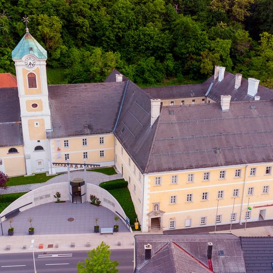 Ein Luftbild eines historischen Gebäudes mit einem Glockenturm, umgeben von Bäumen und einem gepflasterten Bereich. Das Gebäude hat ein graues Dach, mehrere Fenster und einen kleinen Bogengang.