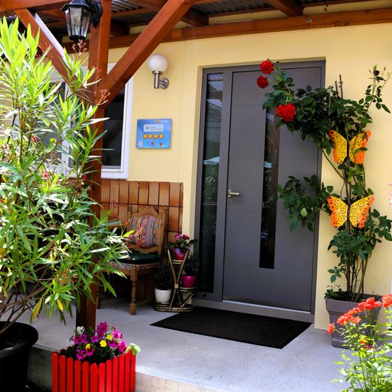 Ein einladender Vorgarten mit einer grauen Tür und roten Blumen, umgeben von Topfpflanzen und einem Holzstuhl. Die Wand hat ein Schild und eine Lampe, und der Boden ist mit einem schwarzen Teppich bedeckt.