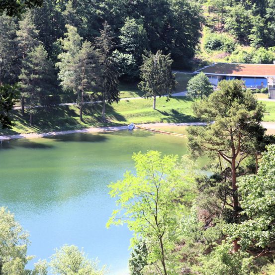 Ein ruhiger See inmitten eines üppigen Waldes mit einem kleinen Boot auf dem Wasser und einem Gebäude in der Nähe.