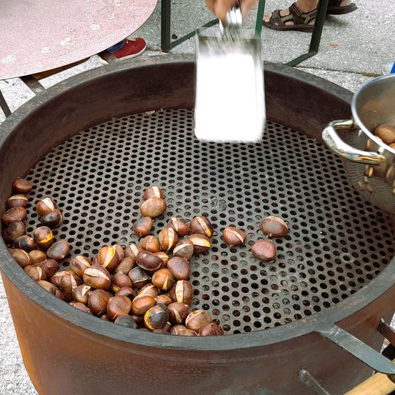 Eine Person fügt gerösteten Kastanien in einem Metallfass mit einem perforierten Gitter Salz hinzu. Ein Edelstahlbehälter steht in der Nähe.