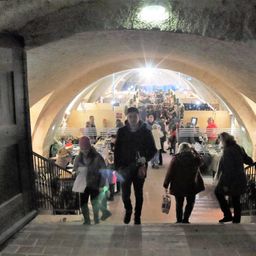 Ein schwach beleuchteter Tunnel mit gewölbten Decken, wo mehrere Menschen gehen. Einige tragen Taschen, und andere gehen in Richtung eines hellen Lichts.