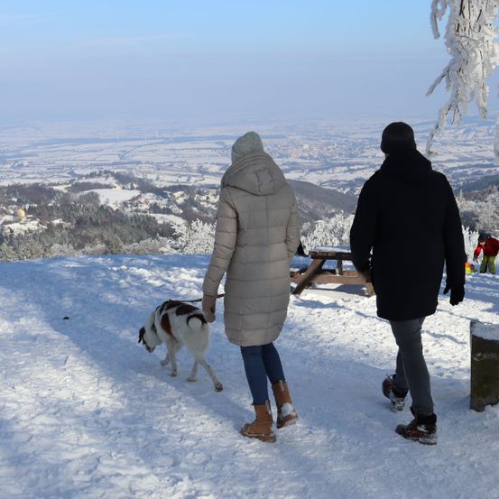 Ein Paar geht mit seinem Hund auf einem verschneiten Berg, mit einem Picknicktisch und einer entfernten Aussicht auf ein Tal.