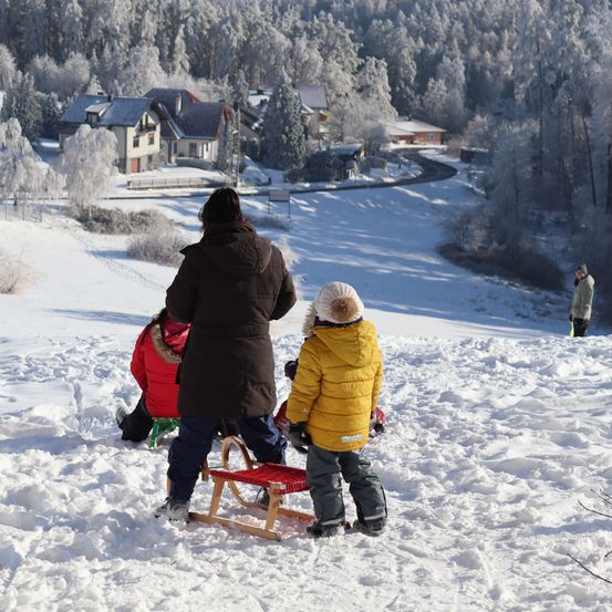 Eine verschneite Landschaft mit einer Gruppe von Menschen, einer mit einem Schlitten, und einem Haus im Hintergrund.