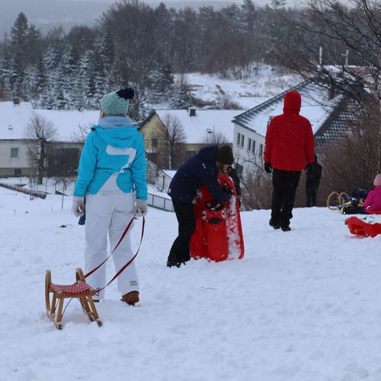 Eine Familie genießt das Schlittenfahren auf einem verschneiten Hügel. Häuser und Bäume sind schneebedeckt. Eine Person hilft einer anderen mit einem roten Schlitten.