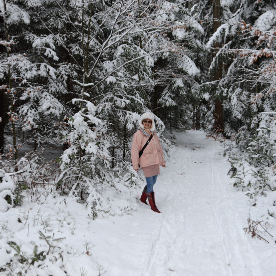 Eine Frau in Winterkleidung steht in einem verschneiten Wald, umgeben von schneebedeckten Bäumen. Sie trägt einen Hut, Sonnenbrille und Stiefel.