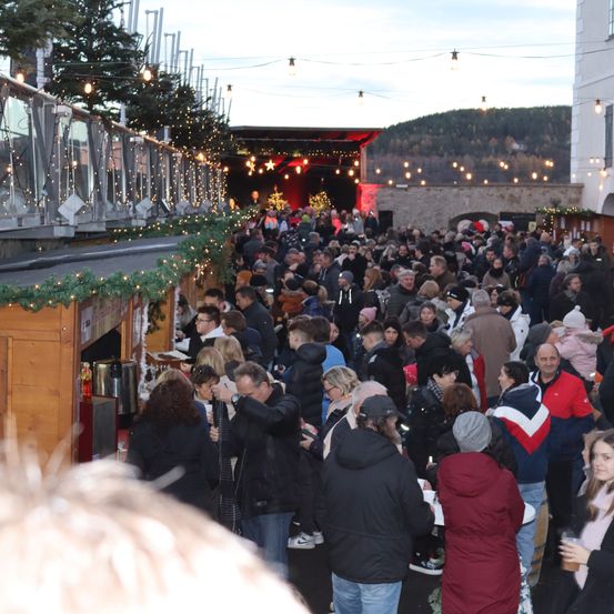 Eine große Menschenmenge versammelt sich auf einem Weihnachtsmarkt. Die Leute stöbern durch Stände mit Dekorationen, und einige machen Fotos. Im Hintergrund befindet sich eine Bühne mit einem Weihnachtsbaum und festlichen Lichtern.