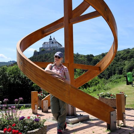 Eine Frau mit Sonnenbrille und Hut lehnt sich an eine hölzerne Spiralstruktur mit einem Schloss im Hintergrund. Die Spirale ist von einem Garten mit Blumen und Bäumen umgeben.