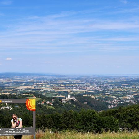Eine Frau steht vor einem Schild auf einem Hügel und fotografiert die Landschaft darunter. Das Tal ist mit Gebäuden gespickt, umgeben von Grün. Der Himmel ist blau mit verstreuten Wolken.