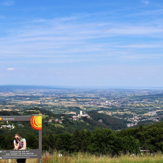 Eine Frau steht vor einem Schild auf einem Hügel und fotografiert die Landschaft darunter. Das Tal ist mit Gebäuden gespickt, umgeben von Grün. Der Himmel ist blau mit verstreuten Wolken.